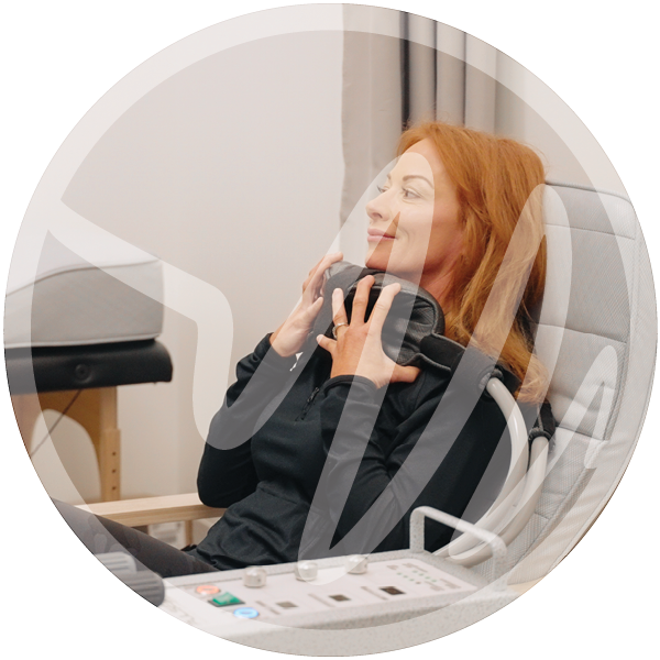 A woman relaxes in a modern therapy chair while using a neck and shoulder massage device, surrounded by clinical wellness equipment in a serene setting.